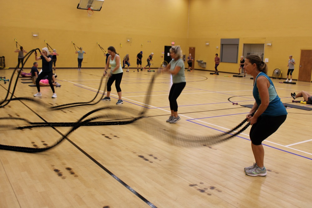 a group of women in the foreground with a group of people in the background exercising. the women in the foreground are swinging large black ropes